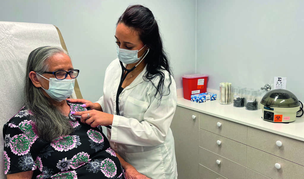 Nurse practitioner listening to and examining patient by listening to her lungs with a stethoscope and gentle has her hand on the patient's shoulder.